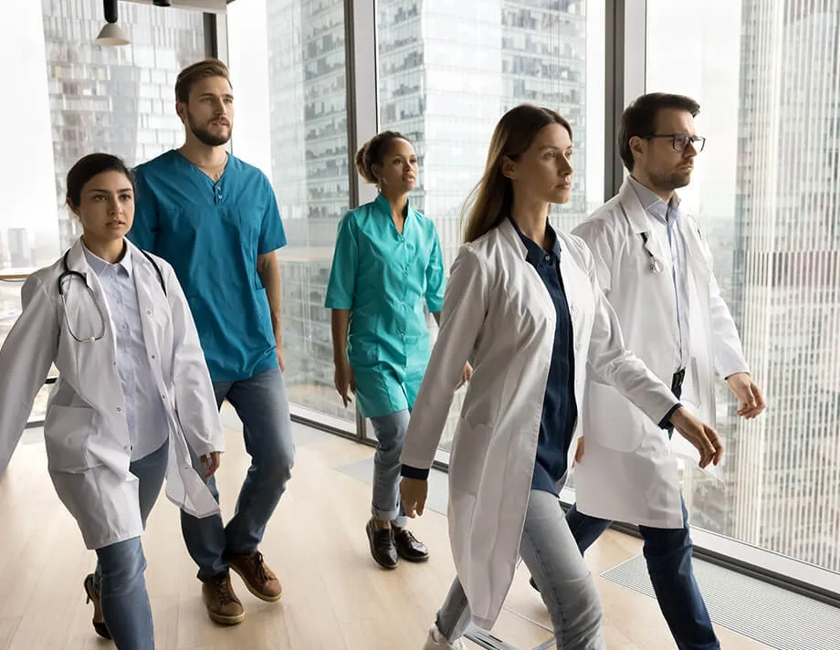A diverse team of medical professionals in lab coats and scrubs walking with confidence in a modern healthcare facility.
