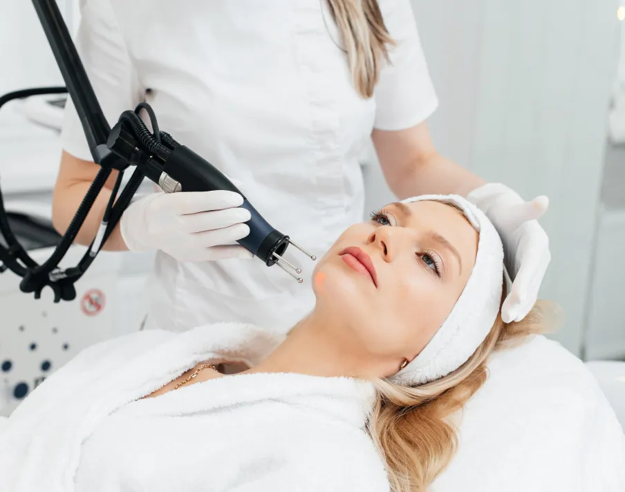 Professional performing light-based facial treatment on woman in dermatology clinic