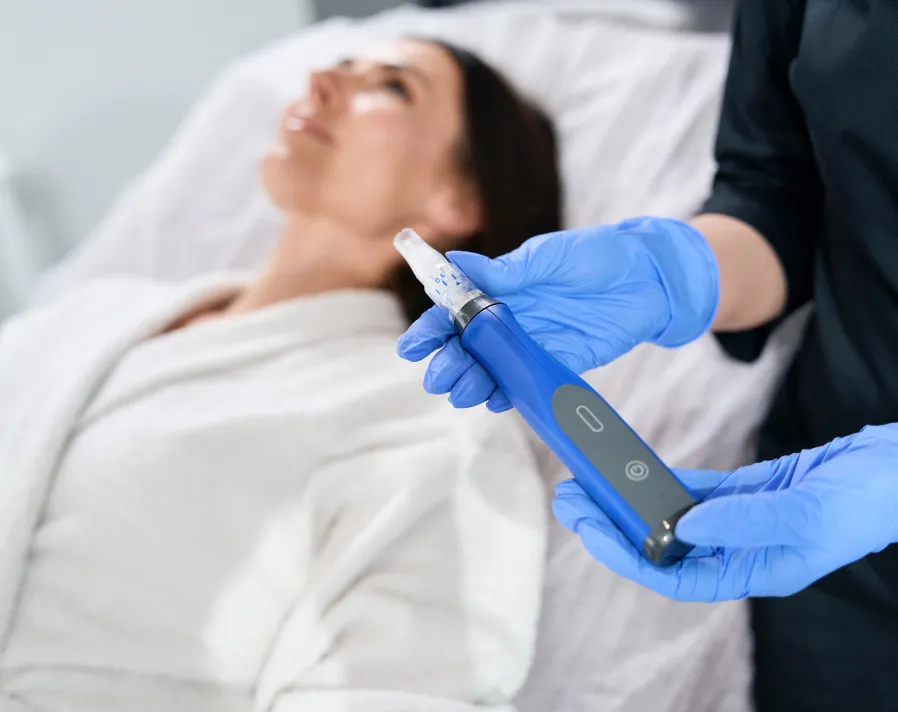 Professional holding microneedling device preparing to treat woman’s face in clinical spa setting